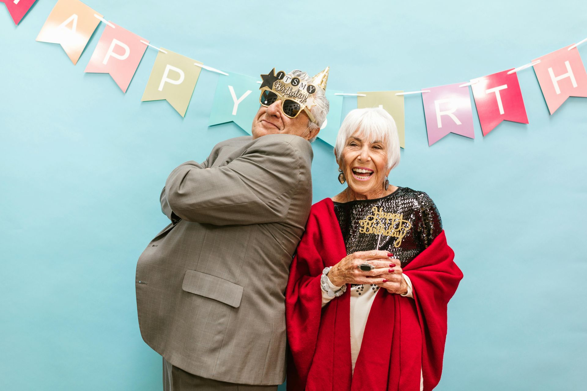 Elderly couple celebrating birthday; man in crown, woman in red, teal backdrop, 